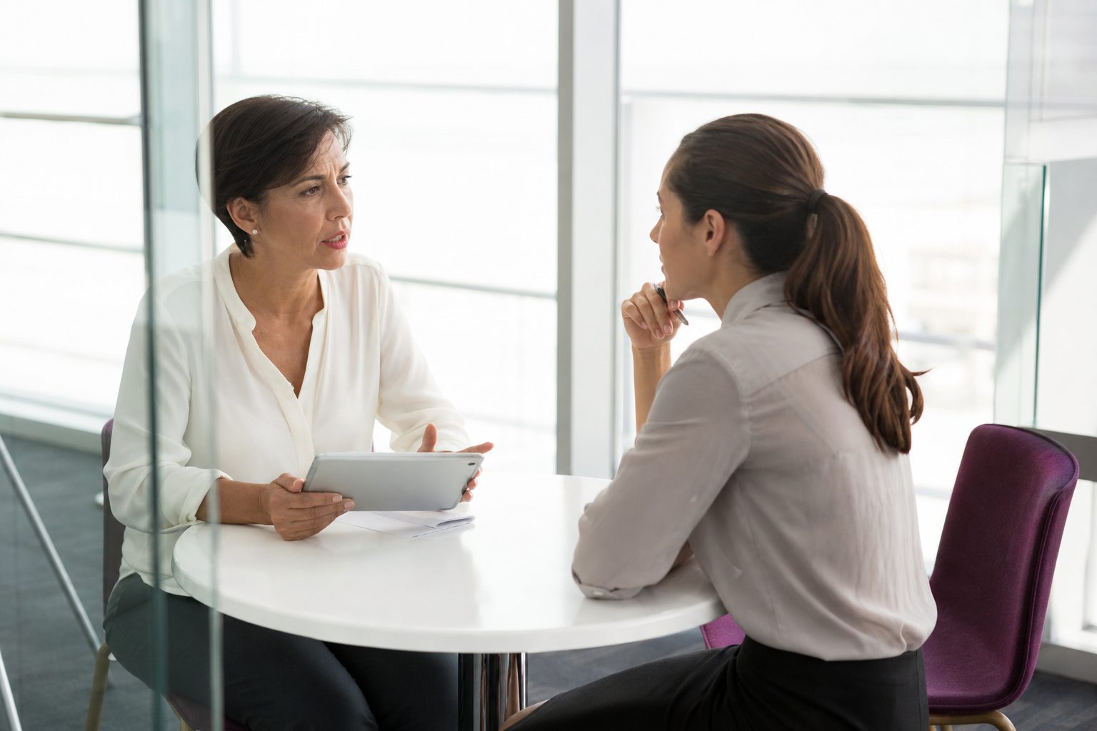 Two colleagues in conversation across a white table in a bright glass meeting room